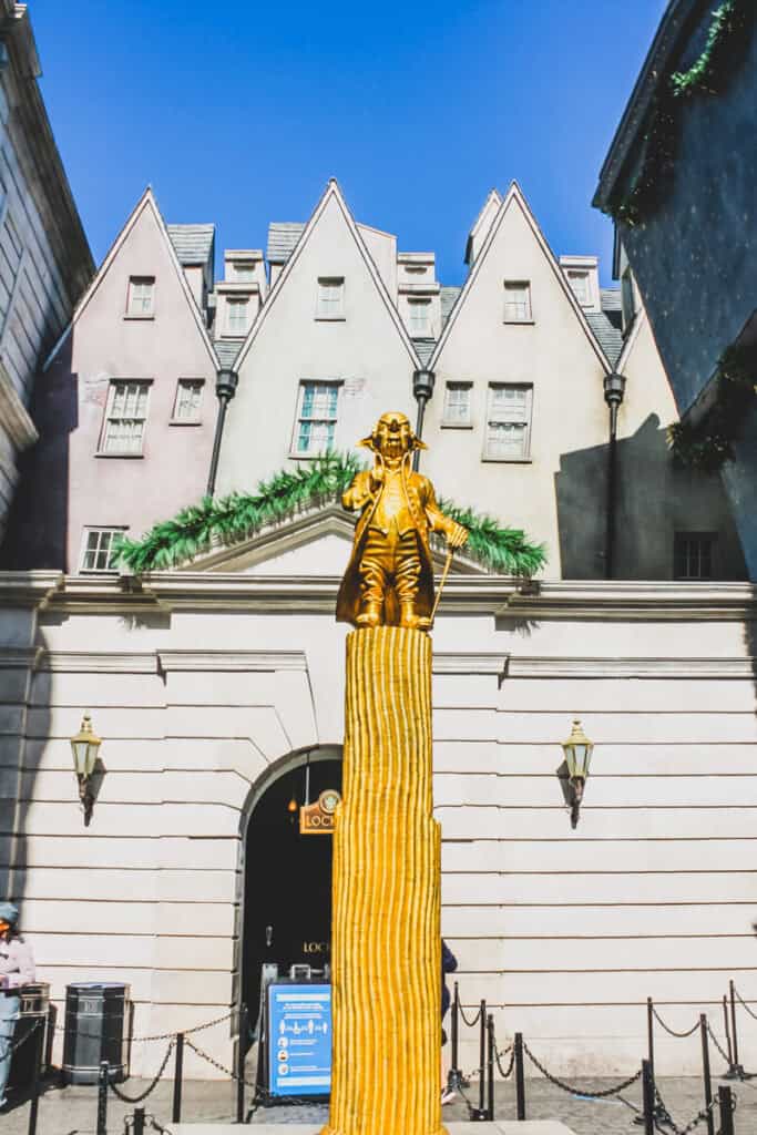 Golden statue of Golbin in front of white building. This is next to the entrance of the Gringotts ride at Harry Potter World
