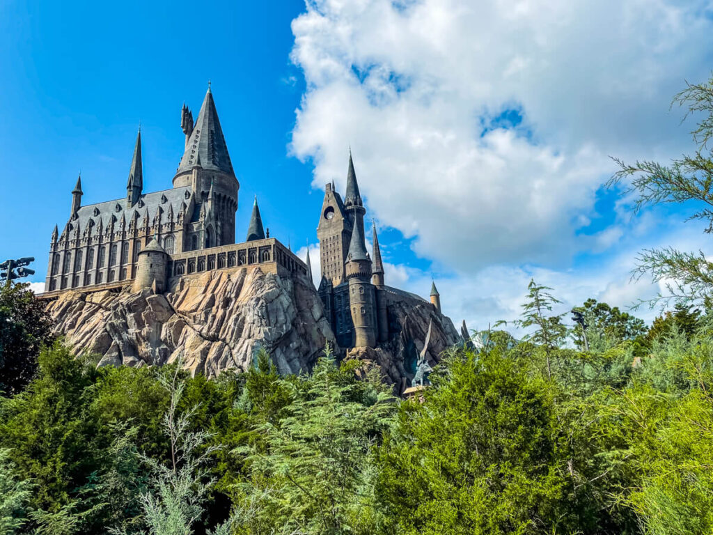 Hogwarts Castle in front of blue sky with clouds and behind greenery. This castle is the main attraction at Harry Potter World