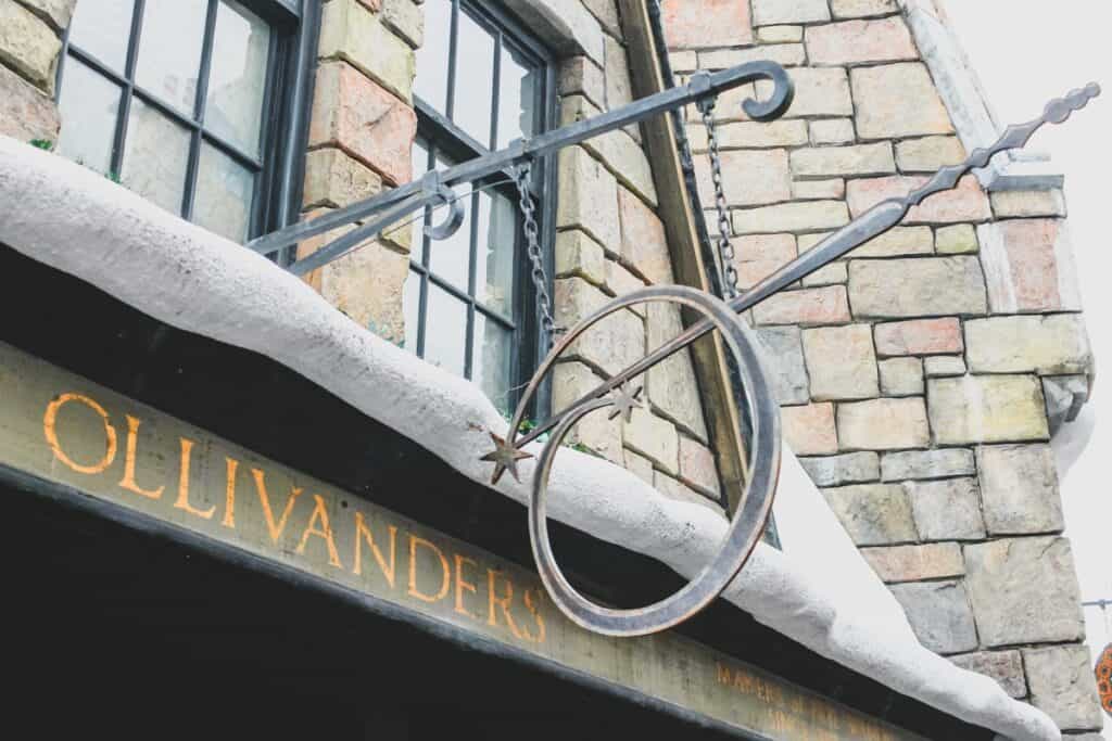metal wand and circle sign in front of snowy building reading Ollivanders. This is the wand shop at Harry Potter World