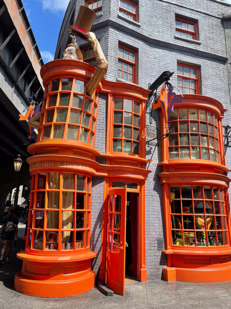 orange window frames along the outside of a building with an animatronic man holding a hat. This is the entrance to Weasley's Wizard Wheezes in Diagon Alley