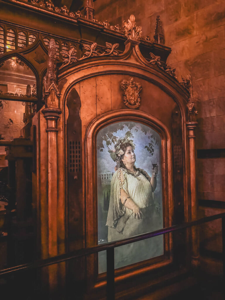 The enchanted painting of the Fat Lady guarding the Gryffindor common room door. She holds a goblet and is framed in an elaborate golden archway with gothic Hogwarts detailing. Located inside the queue for Forbidden Journey ride at Harry Potter World