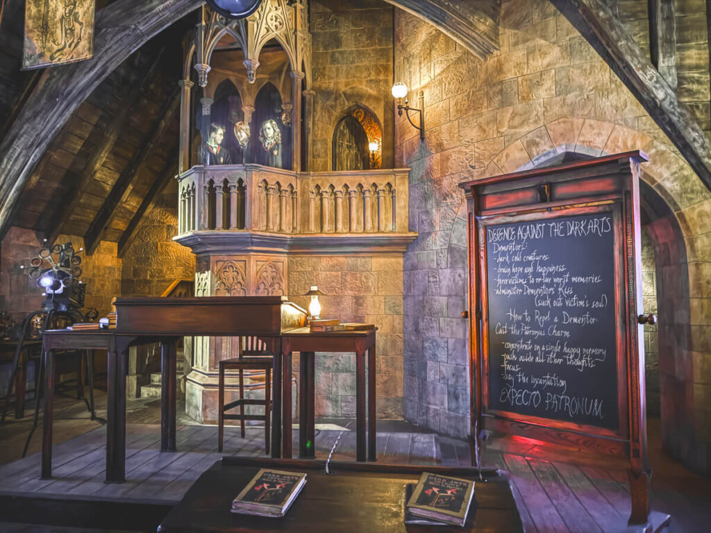 A classroom set with dark wood furniture and a chalkboard covered in instructions on repelling Dementors. A projection of Harry, Hermione, and Ron appears on a stone balcony in the background. This is one of the rooms of the queue for the Forbidden Journey ride at Harry Potter World