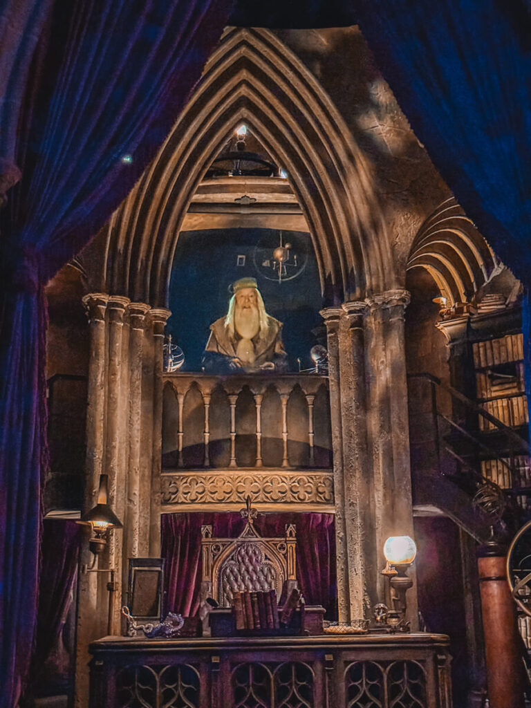 Inside the Forbidden Journey ride queue, a projection of Dumbledore appears behind a Gothic-style stone archway with rich purple curtains framing the scene. The setting is detailed with his desk, books, and glowing lamps, creating a magical atmosphere straight from the films.