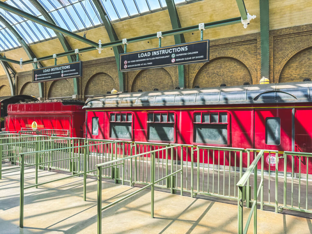 A bright red Hogwarts Express train is parked under a glass canopy in King’s Cross Station at Harry Potter World Diagon Alley. Loading instructions are displayed above, and the queue is empty, ready for riders.