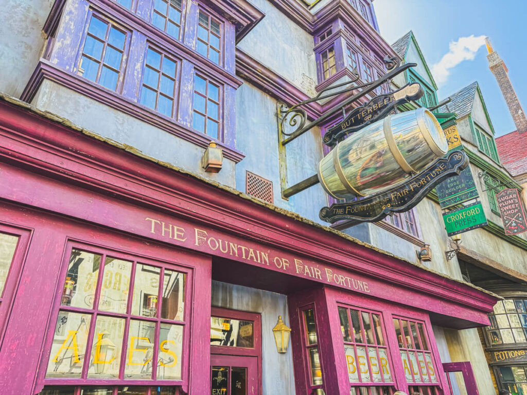 A vibrant exterior of The Fountain of Fair Fortune pub in Diagon Alley, featuring a shiny silver butterbeer barrel sign. The facade is painted deep purple and red with whimsical shop signs nearby.
