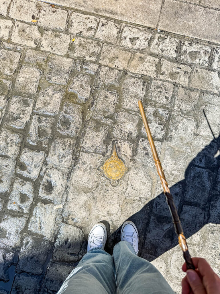 A pair of sneakers and jeans stand next to a golden medallion embedded in cobblestone, indicating an interactive wand spot in Diagon Alley. A hand is holding a Harry Potter-style wand mid-spell, casting magic on the street.