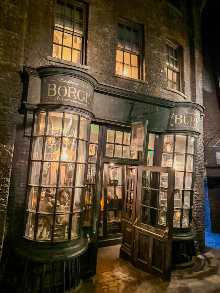 The storefront of Borgin and Burkes in Knockturn Alley in Harry Potter World glows ominously under warm lighting. The shop windows are filled with eerie magical artifacts, capturing the dark, mysterious vibe of this hidden alley.