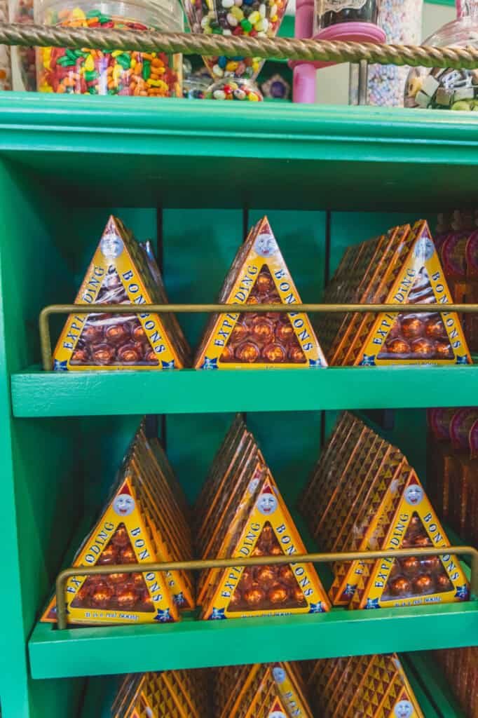 A shelf inside Honeydukes at Harry Potter World displays rows of triangular boxes of Exploding Bonbons, with shiny orange treats arranged behind bright yellow packaging. These fiery sweets are a fun and flavorful favorite among visitors to the Wizarding World.