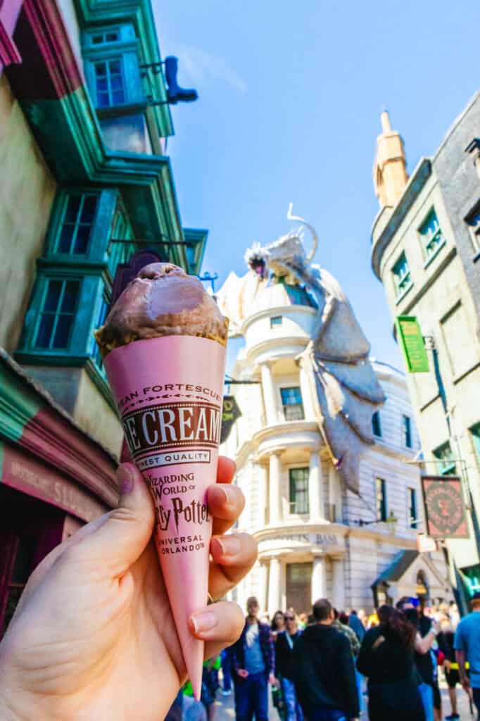 A hand holds a cone of chocolate ice cream from Florean Fortescue’s Ice Cream Parlour in Diagon Alley, with the fire-breathing dragon atop Gringotts Bank towering in the background. This magical snack stop is a fan-favorite photo op and a delicious highlight of any visit to Harry Potter World.