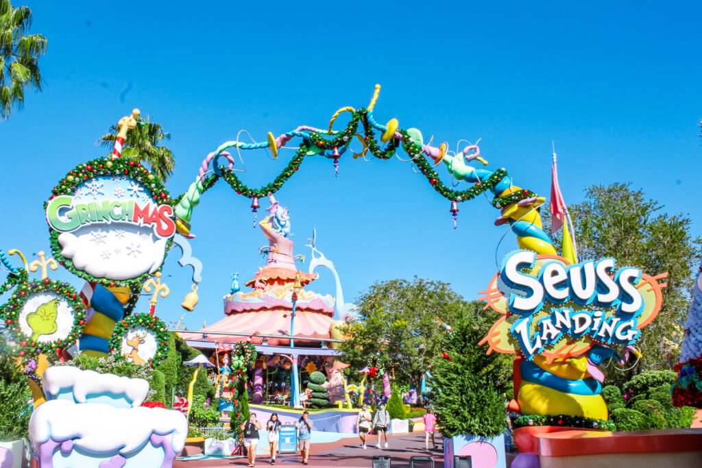 Entrance to Seuss Landing at Universal Orlando, decorated for the holidays with colorful garlands and ornaments. A whimsical arch connects two signs: one reads “Grinchmas” with snowy accents, and the other reads “Seuss Landing,” surrounded by Dr. Seuss-style swirls and Christmas decorations, with the area filled with vibrant colors and playful structures.