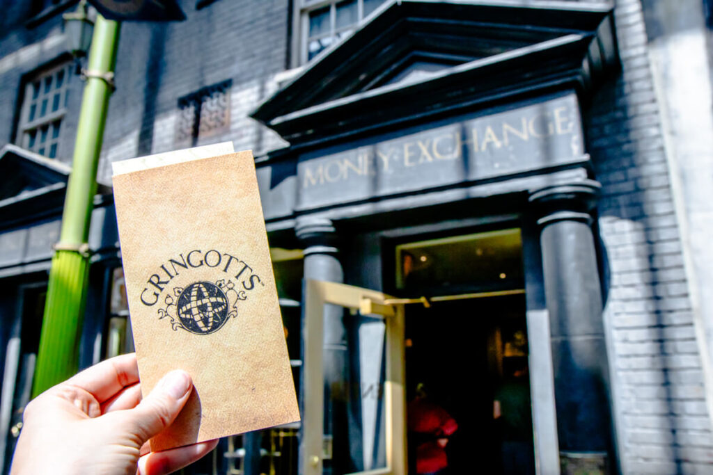 A hand holds up a Gringotts-branded envelope in front of the Money Exchange building at the Wizarding World of Harry Potter in Universal Studios Florida. Exchanging Muggle money for wizarding currency is a fun, immersive experience to include in any Harry Potter World itinerary.