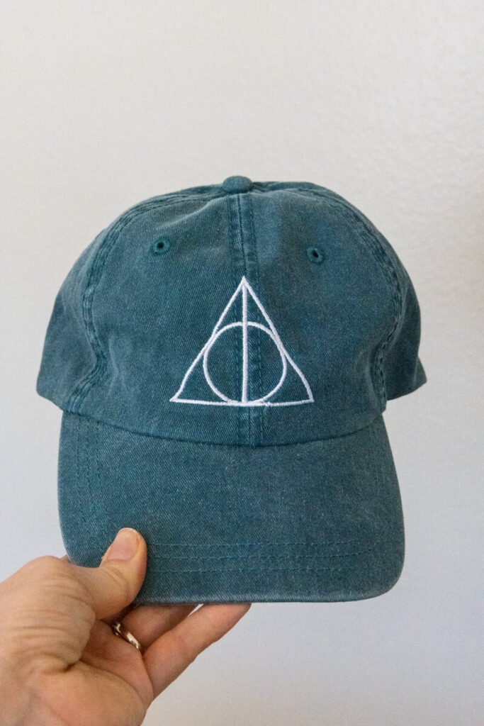 hand holding a blue baseball cap with the Deathly Hallows logo