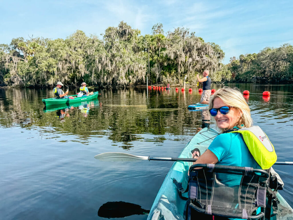 A woman in a kayak smiles back at the camera, with several manatees in the water and other kayakers in front of her, in front of the buoy line at Blue Spring State Park