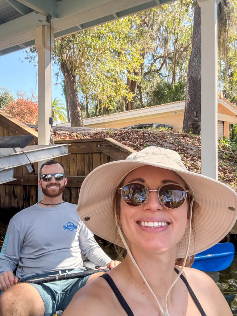 Smiling couple kayaking through the canals of Winter Park Florida with the woman wearing a sun hat and sunglasses while taking a selfie from the front of the kayak.