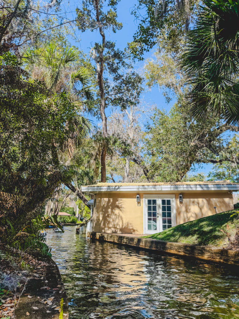 Narrow canal in Winter Park Florida lined with tropical plants and tall trees with Spanish moss beside a lakeside boathouse along the water.