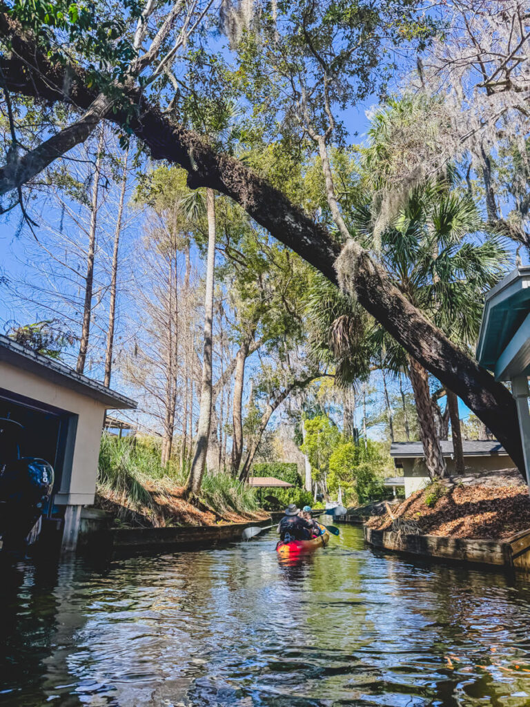 Two people kayaking through a shaded canal in Winter Park Florida beneath arching oak trees and palms with homes and boat houses along the waterway.