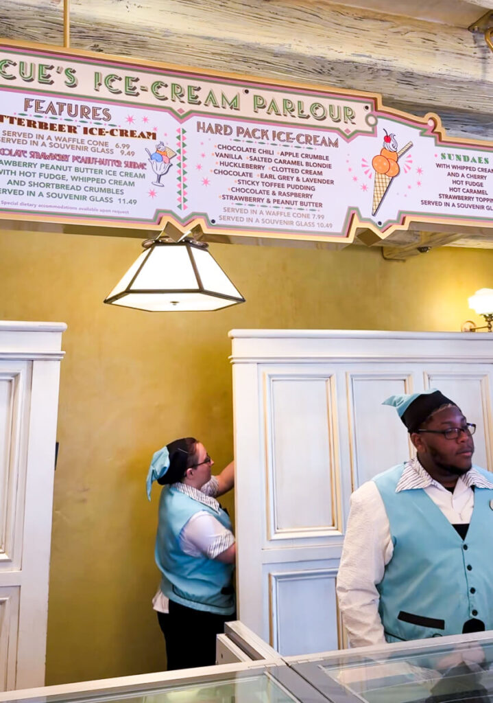 two workers in uniform standing inside Florean Fortescue's ice cream parlor with menu above them