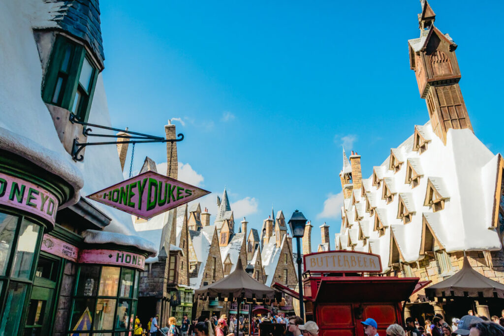 view of Hogsmeade village in Islands of Adventure with Honeydukes sign, butter beer cart, snowy roofs, and blue sky in the background