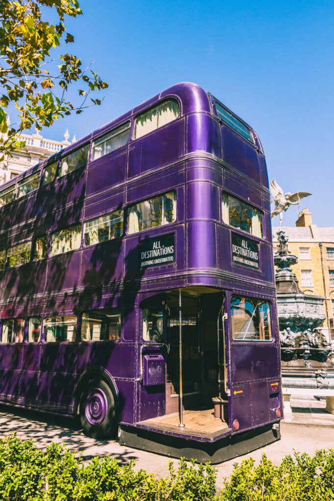 view of the back of the purple triple decker knight bus inside of Universal Studios, with blue skies and green foliage around it