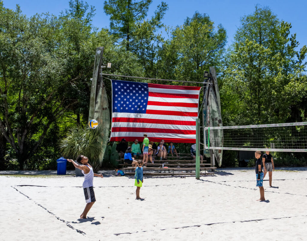 Kids playing beach volleyball on the sand court at Wekiva Island, with a large American flag and bleachers behind them