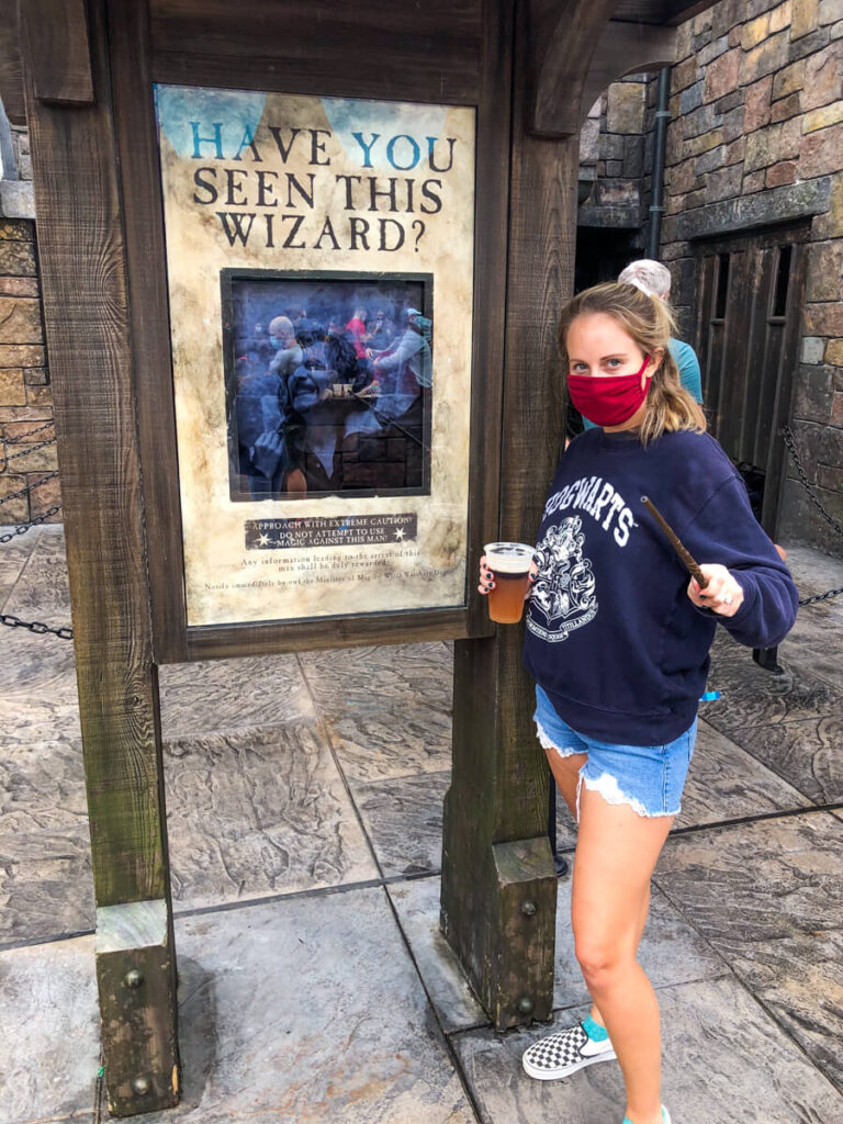 girl in Hogwarts sweater and jean shorts standing in front of Wanted sign for Sirius Black, holding a wand and staring at camera