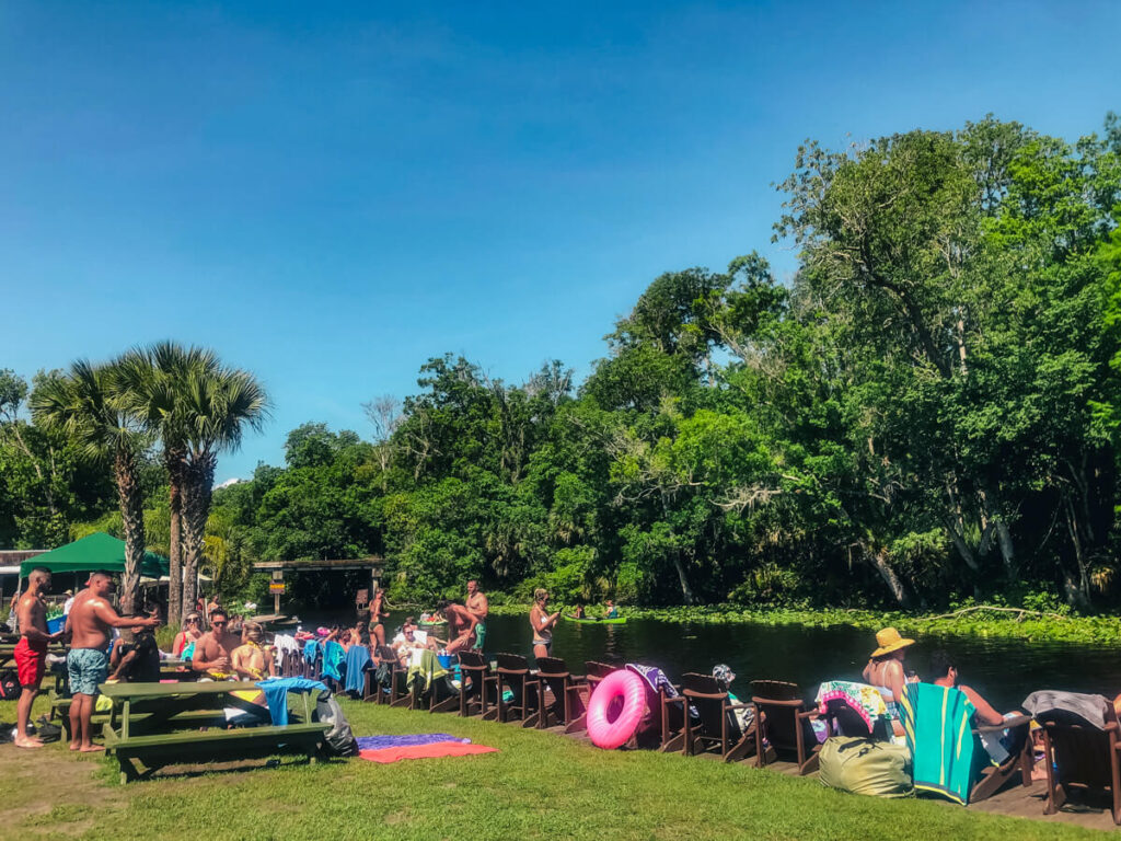 Crowd relaxing in Adirondack chairs and at picnic tables along the riverbank at Wekiva Island on a sunny day